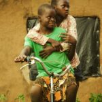 Two African children enjoy a playful moment, sharing a bicycle ride in a rural setting.