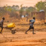 Two children transporting water containers on a wheelbarrow in rural Adjumani, Uganda.