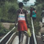 Back view of a girl walking along a railway track carrying a plastic bag, surrounded by greenery.